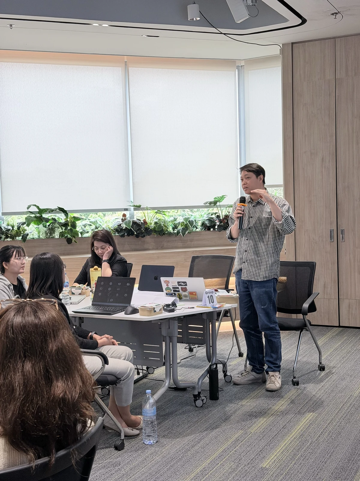 A facilitator speaking beside participants practising with laptops at a workshop table.